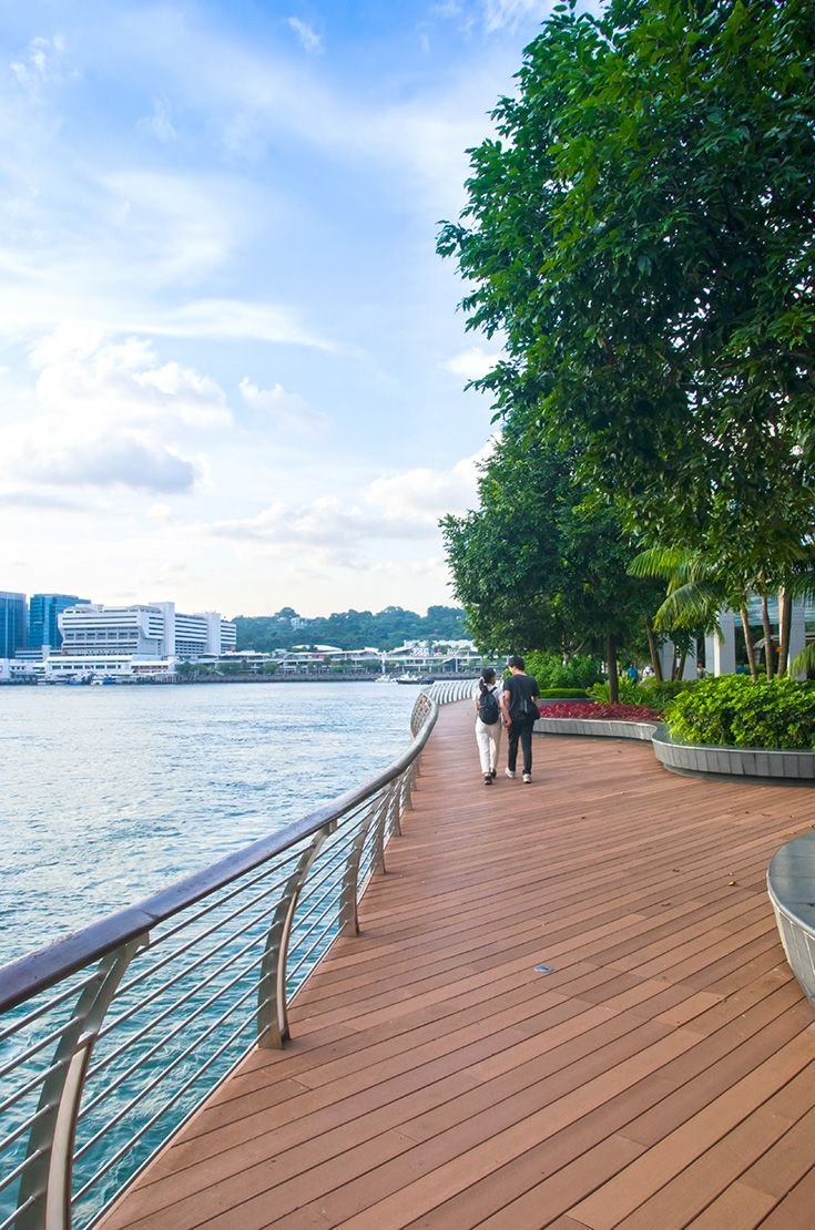 This may contain: two people walking on a boardwalk next to the water with trees and buildings in the background