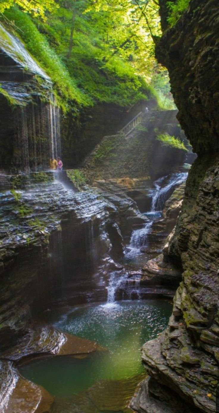 This may contain: a man standing at the base of a waterfall in a canyon surrounded by green trees