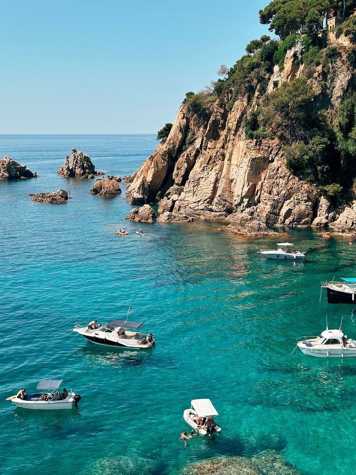 This may contain: several small boats floating in clear blue water near the shore on a rocky cliff face