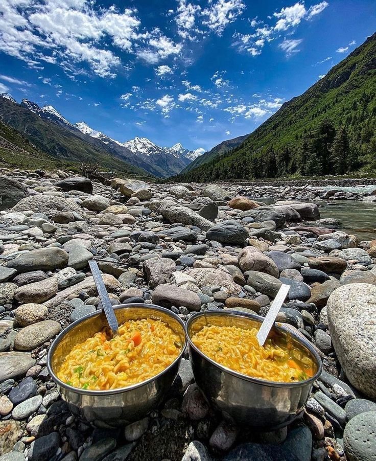 This may contain: two metal bowls filled with food sitting on top of a rocky river bank next to mountains