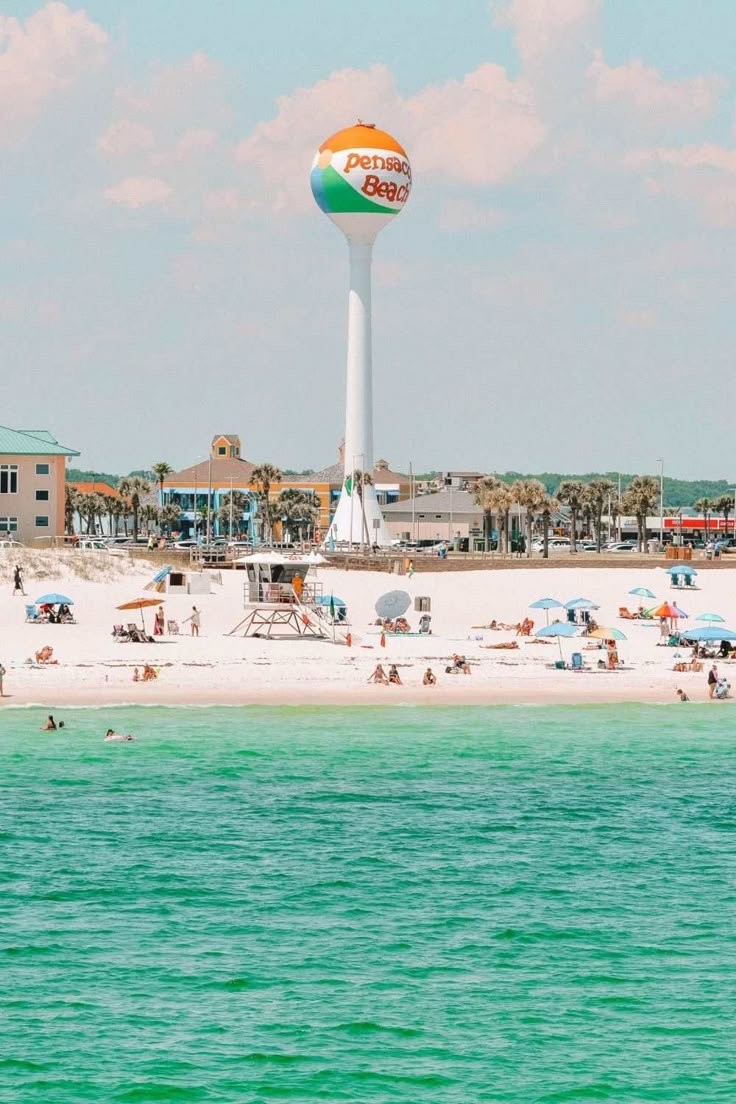 This may contain: the beach is crowded with people and umbrellas under a water tower in the distance