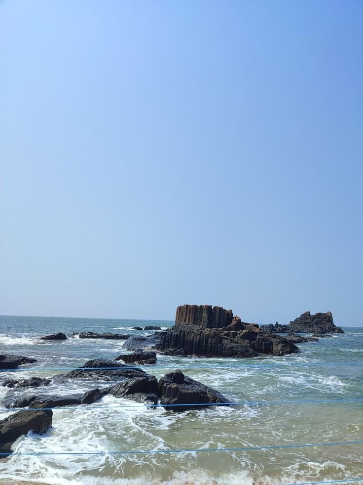 This contains: there is a blue sea with a clear blue sky backdrop. a few geological formations of columnar rhyoltic lava are visible