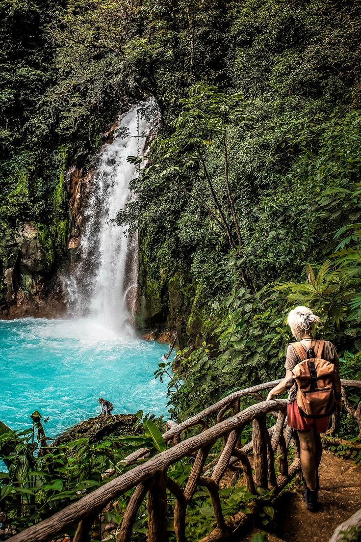 This may contain: a person standing on a bridge looking at a waterfall in the forest with blue water