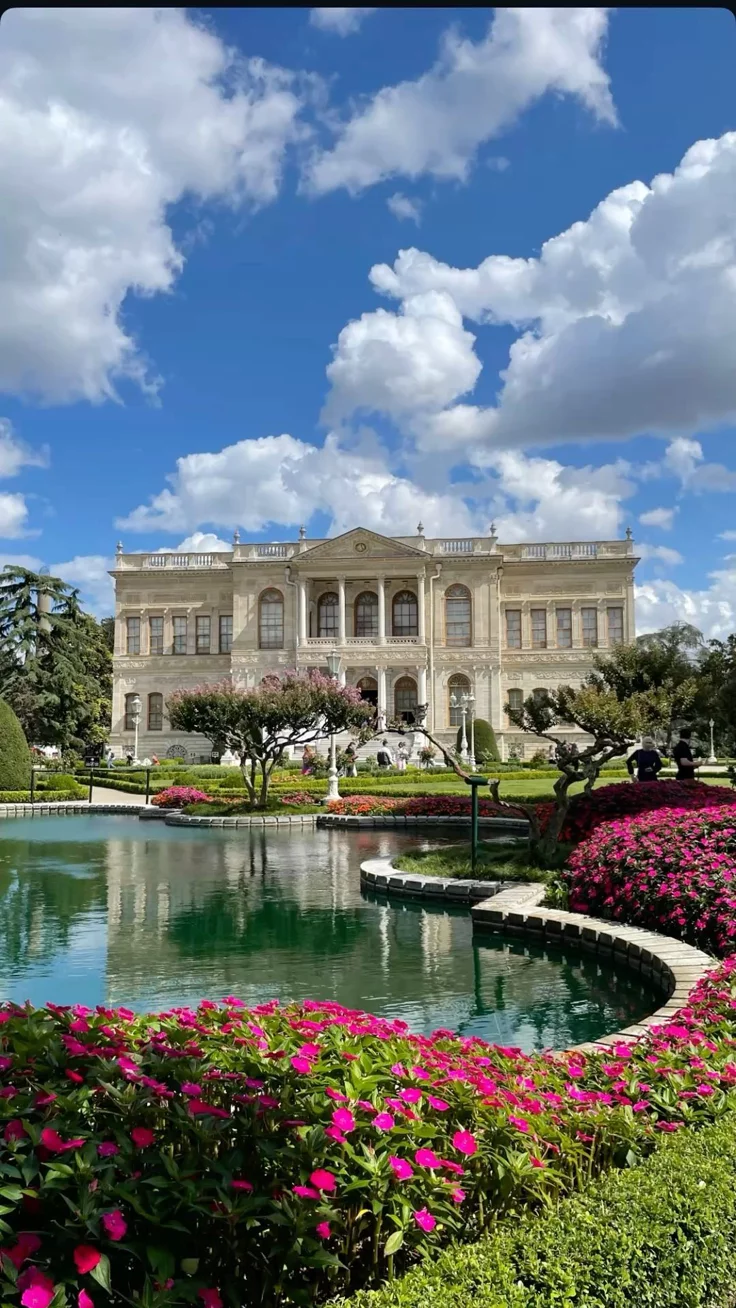 This may contain: a large building with lots of flowers in front of it and a pond on the side