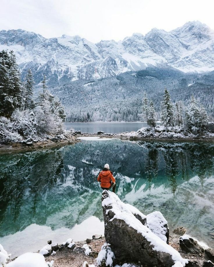 This may contain: a man sitting on top of a rock next to a lake filled with snow covered mountains