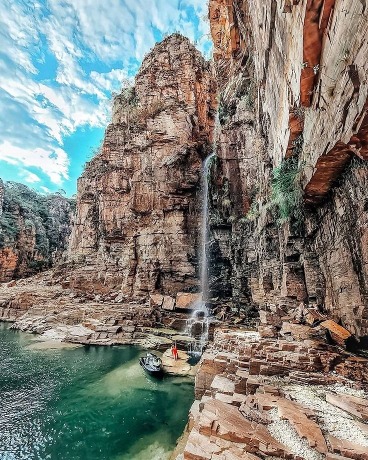 This may contain: a boat is in the water near a waterfall and some rocks with a man standing on it