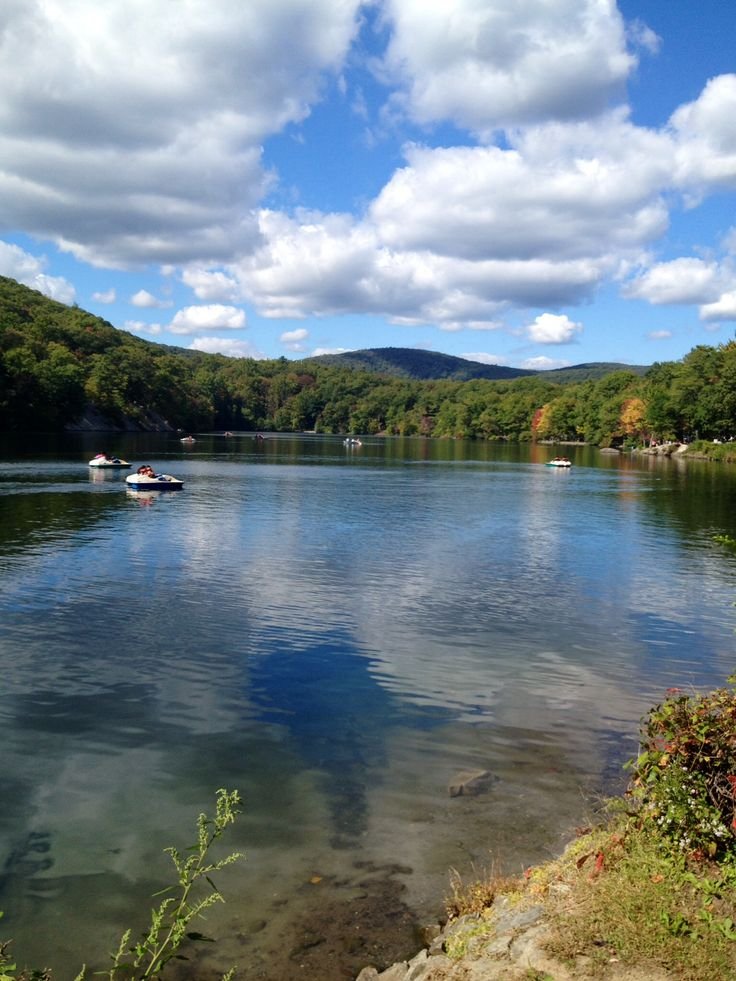 This may contain: a lake surrounded by trees and mountains under a cloudy blue sky with boats in the water