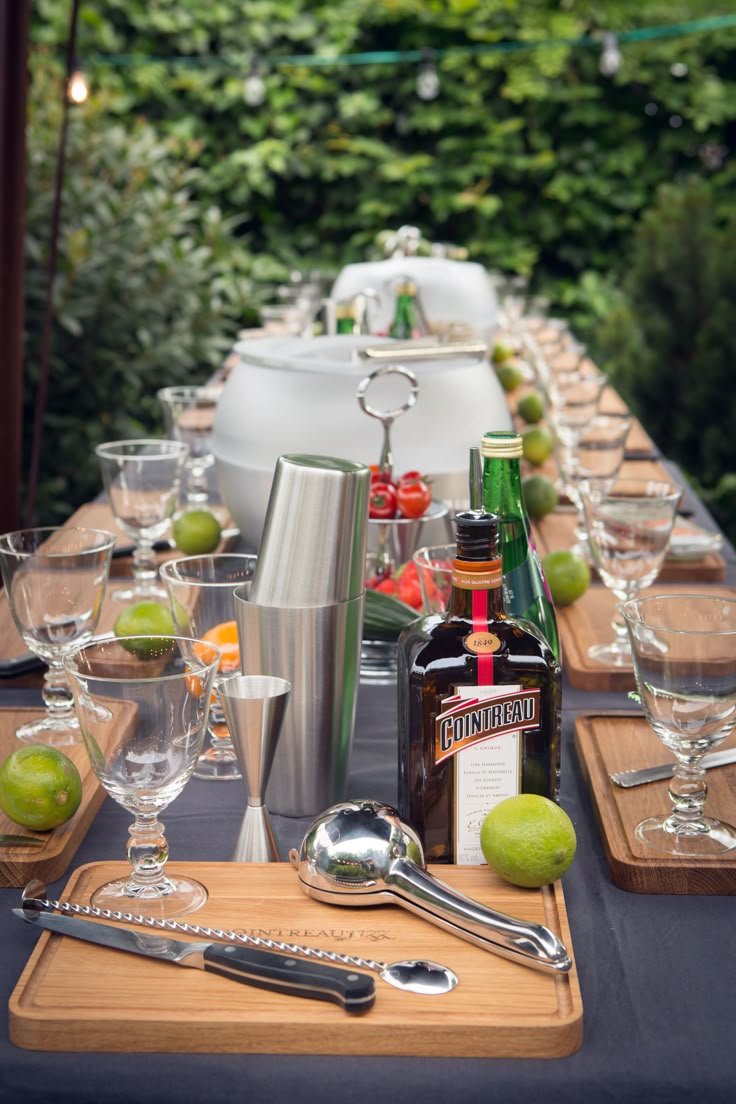 This may contain: a long table is set with glasses, bottles and utensils for an outdoor party