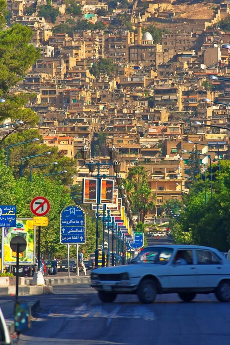 This may contain: a car driving down the road in front of a hill with lots of buildings on it