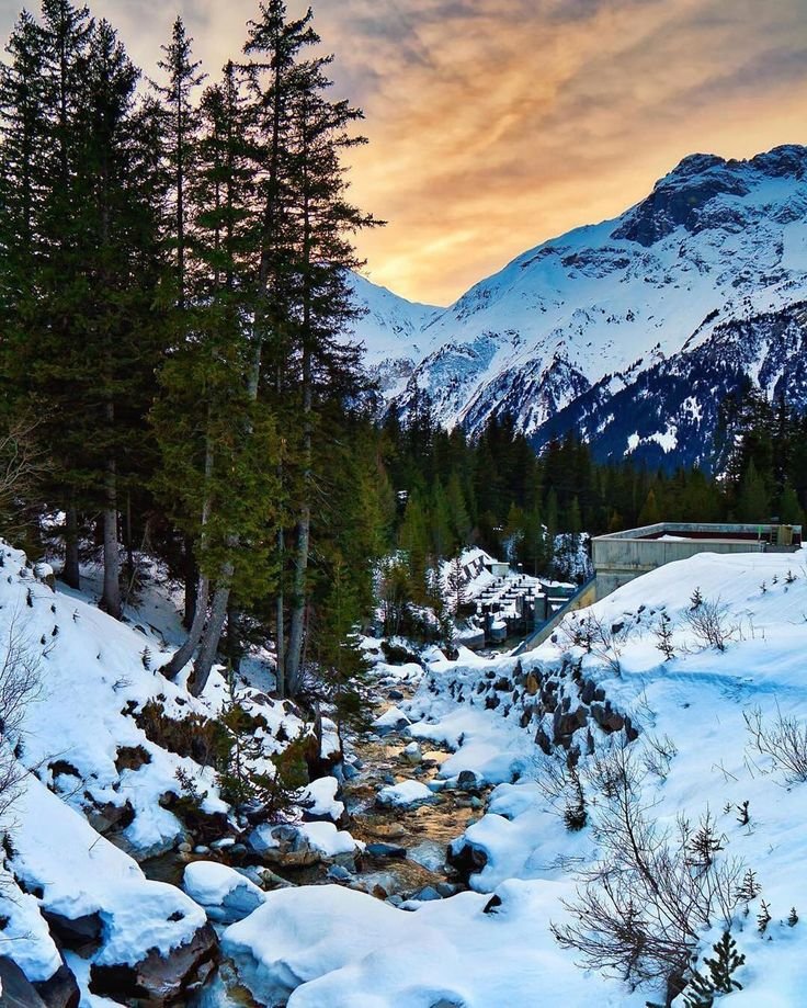 This may contain: a stream running through the middle of a snow covered mountain valley with pine trees and mountains in the background