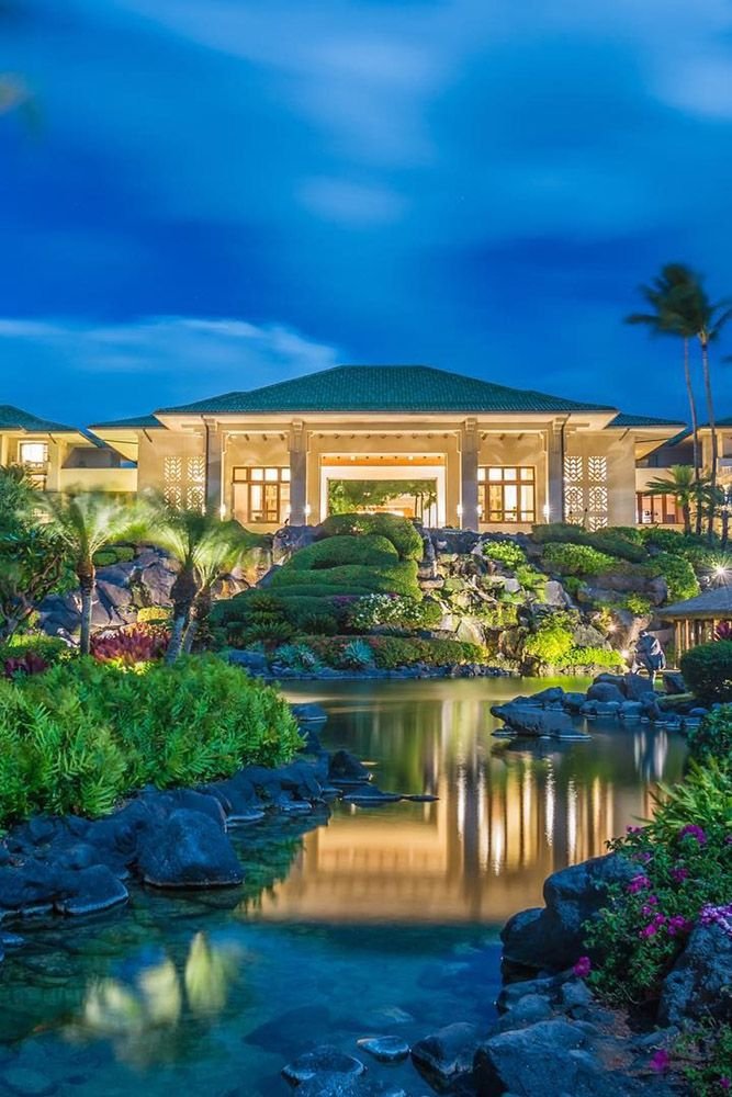 This may contain: a large building sitting on top of a lush green hillside next to a lake at night
