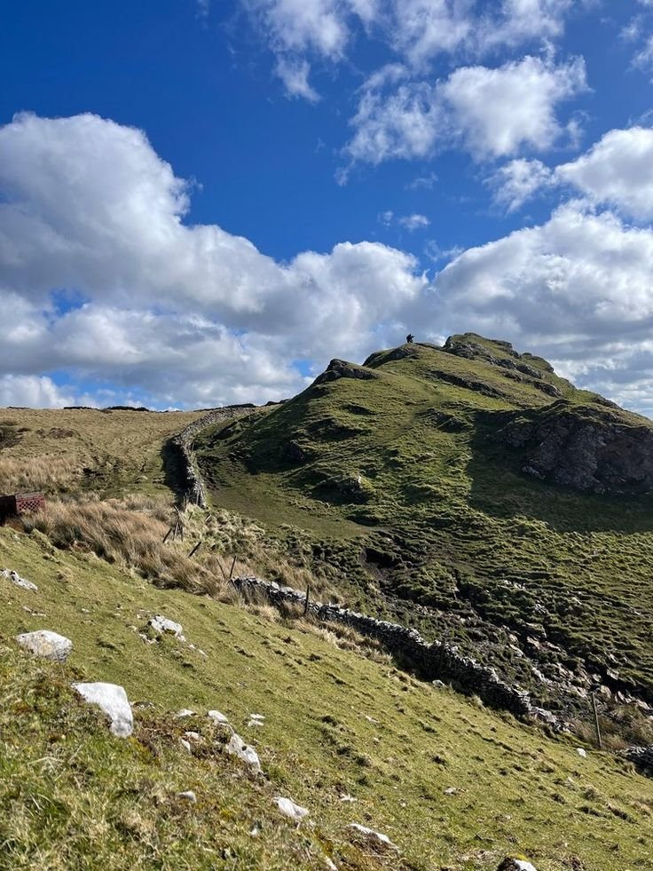 This may contain: a grassy hill with rocks and grass on the side under a blue sky filled with clouds