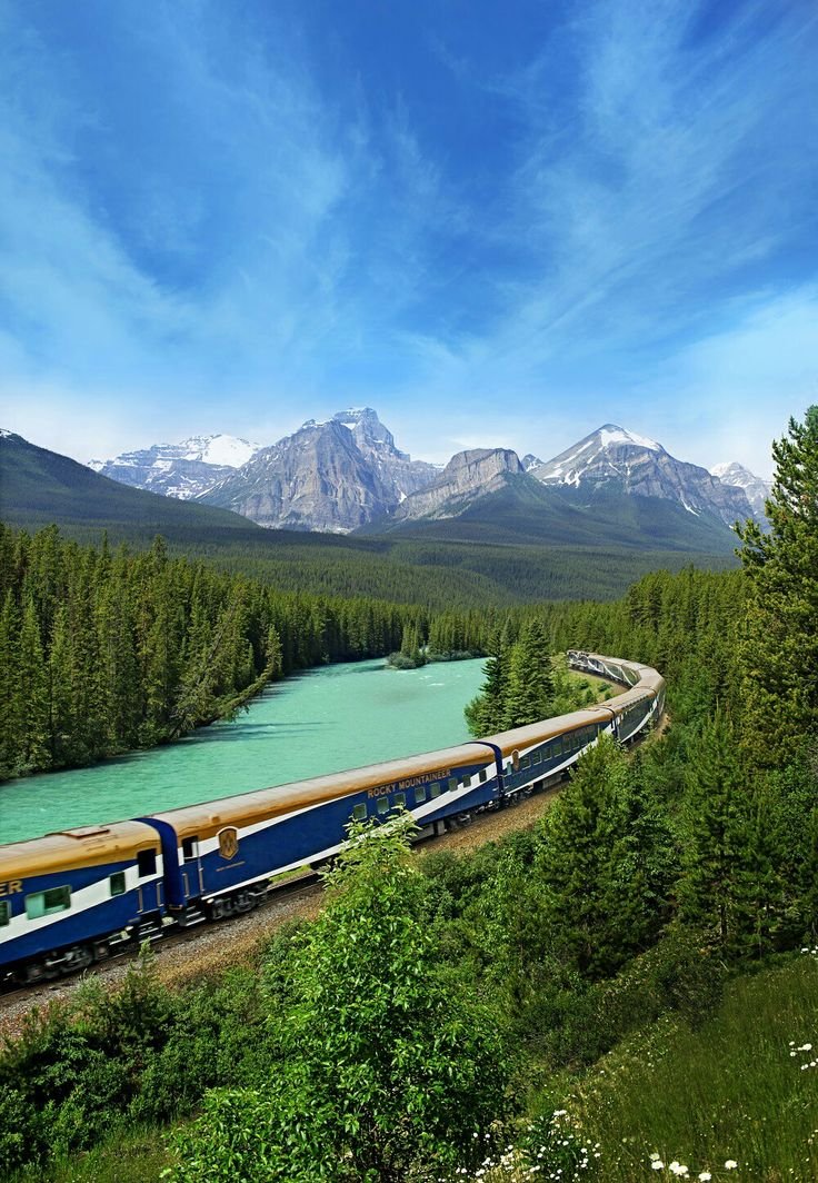 This may contain: a train traveling through the mountains next to a body of water with trees on both sides