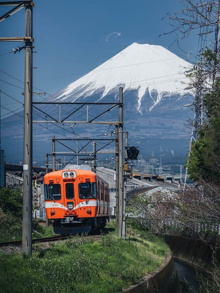 This may contain: an orange and white train traveling past a snow - capped mountain in the background,