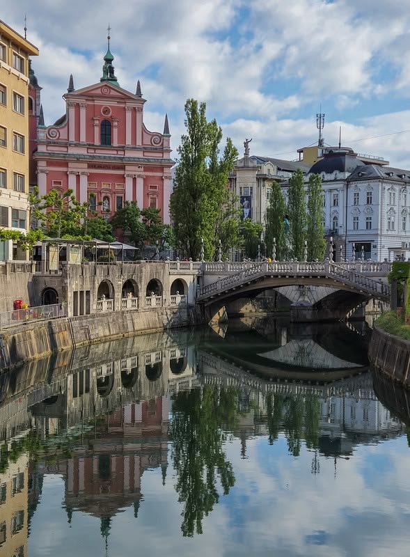 This may contain: a bridge over a river with buildings in the background and water reflecting it's surface