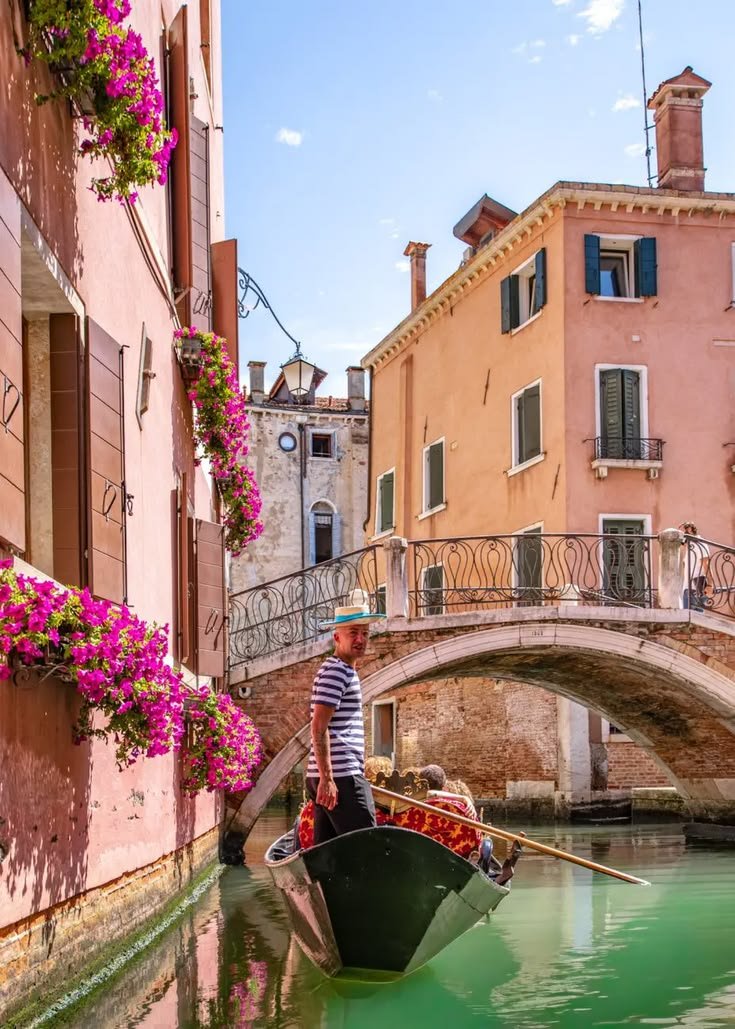This may contain: a man riding on the back of a boat down a river next to tall buildings