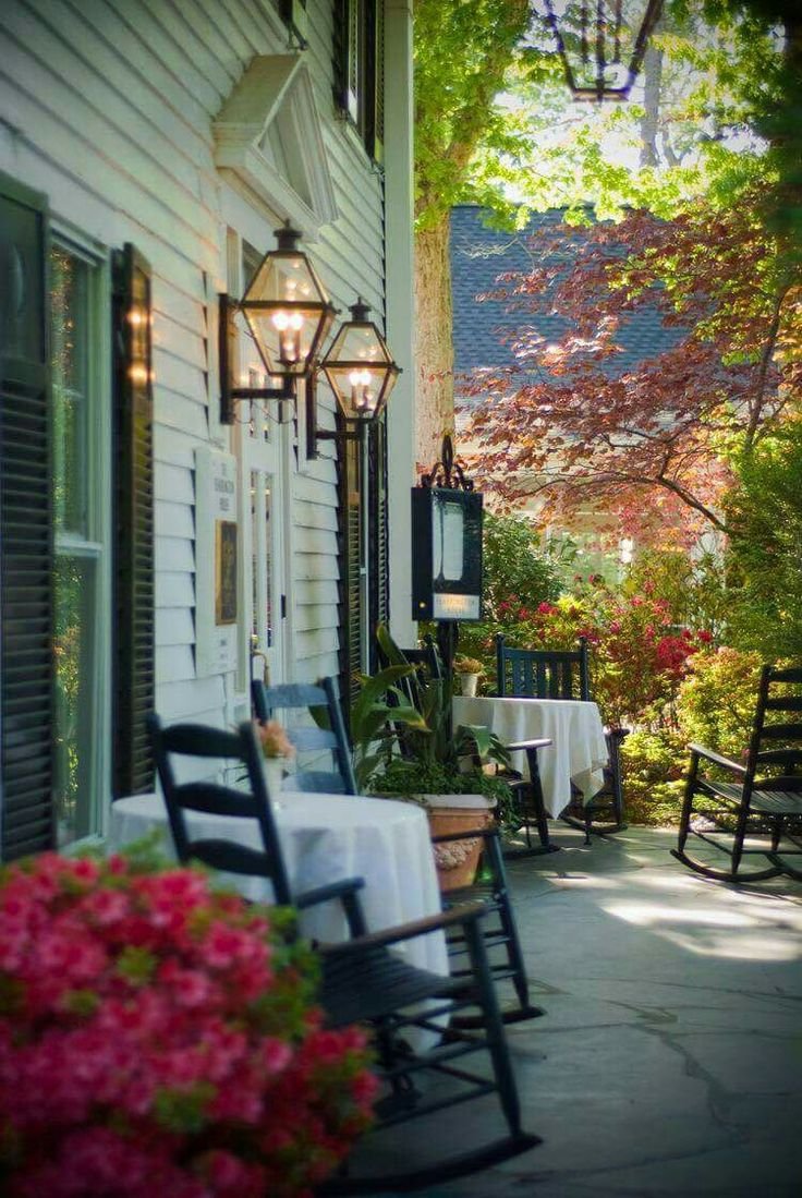 This may contain: a porch with rocking chairs and tables on the side of it next to some flowers