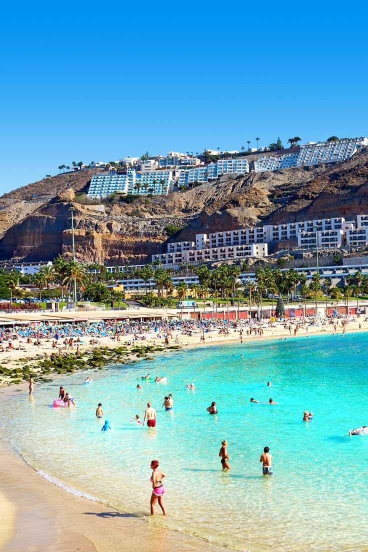 This contains: Playa Amadores in Gran Canaria. Zandstrand met spelende kinderen en op de achtergrond een berg met verschillende hotels.