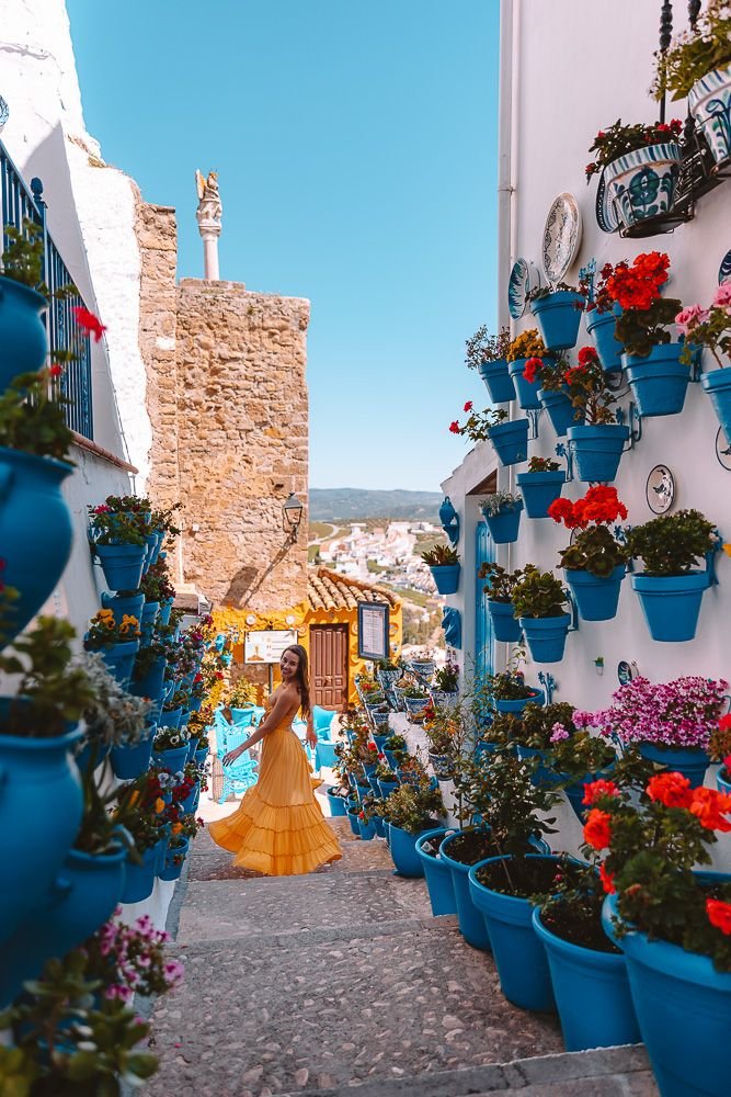 This may contain: a woman in a yellow dress is walking down an alley way with potted plants