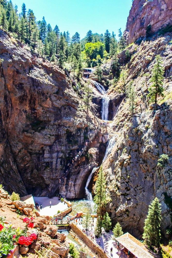 This may contain: a waterfall in the middle of a canyon surrounded by trees and flowers with people walking around it