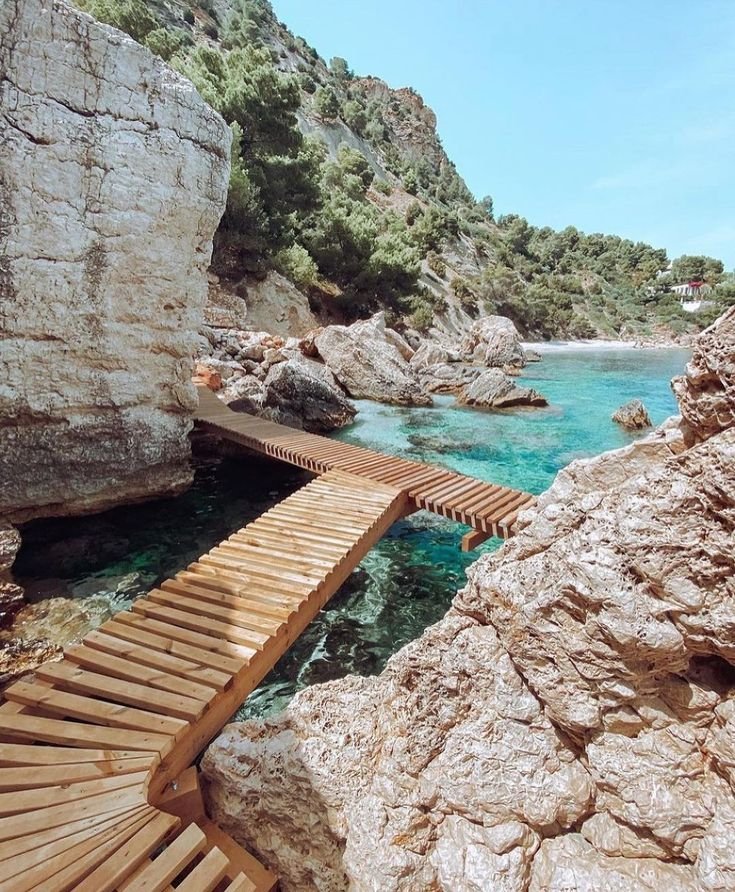This may contain: a wooden walkway over the water leading to a beach area with large rocks and cliffs