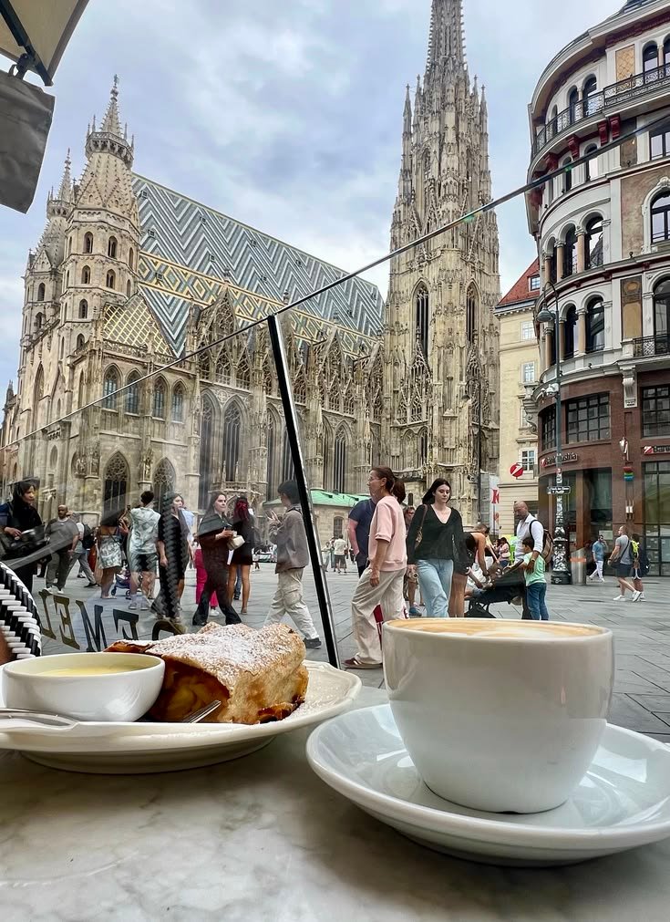This may contain: two cups of coffee on a table in front of a cathedral with people walking around