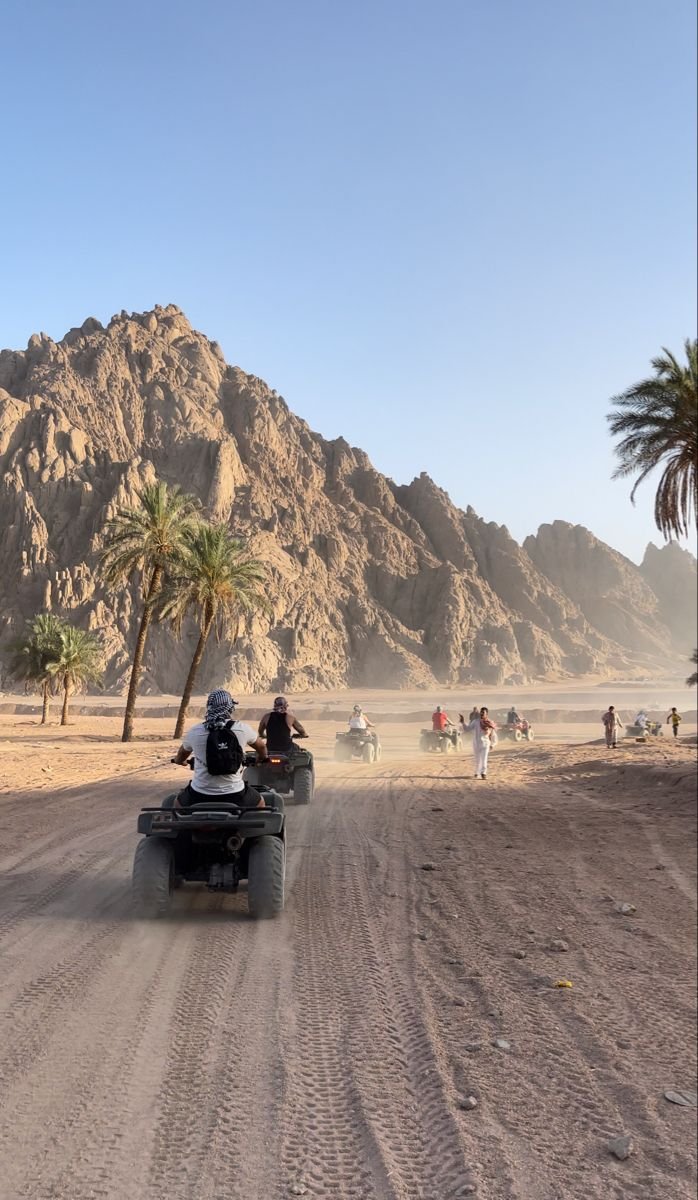 This may contain: two people on four wheelers driving down the road in front of some palm trees