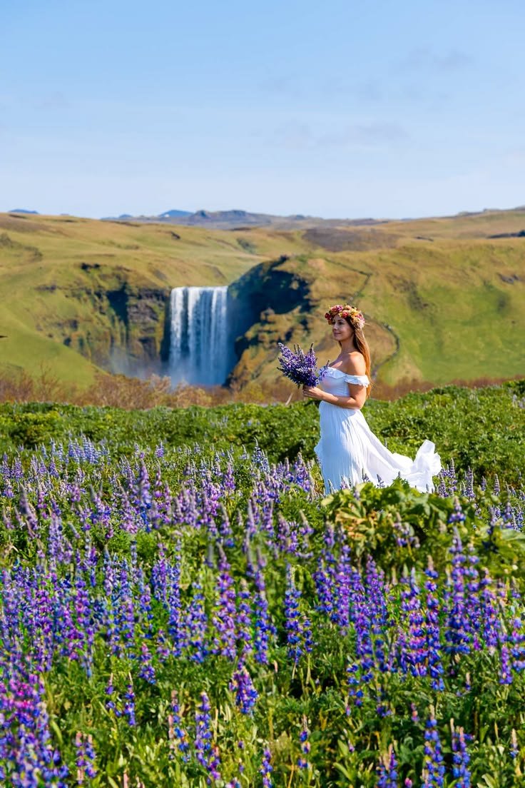 This may contain: a woman in a white dress is walking through a field with purple flowers near a waterfall