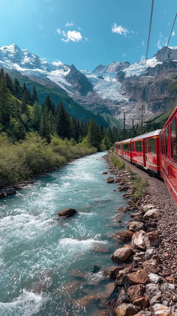 This may contain: a red train traveling down tracks next to a river and mountain covered with snow capped mountains