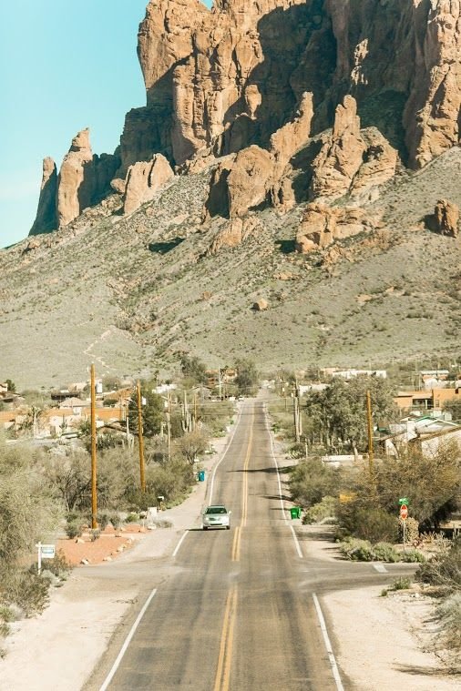This may contain: an empty road in the desert with mountains in the backgrouund and houses on either side