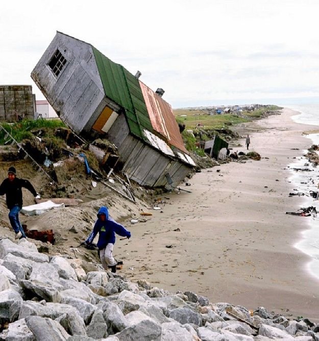 This may contain: two people are walking on the beach near a house that has been blown over by waves