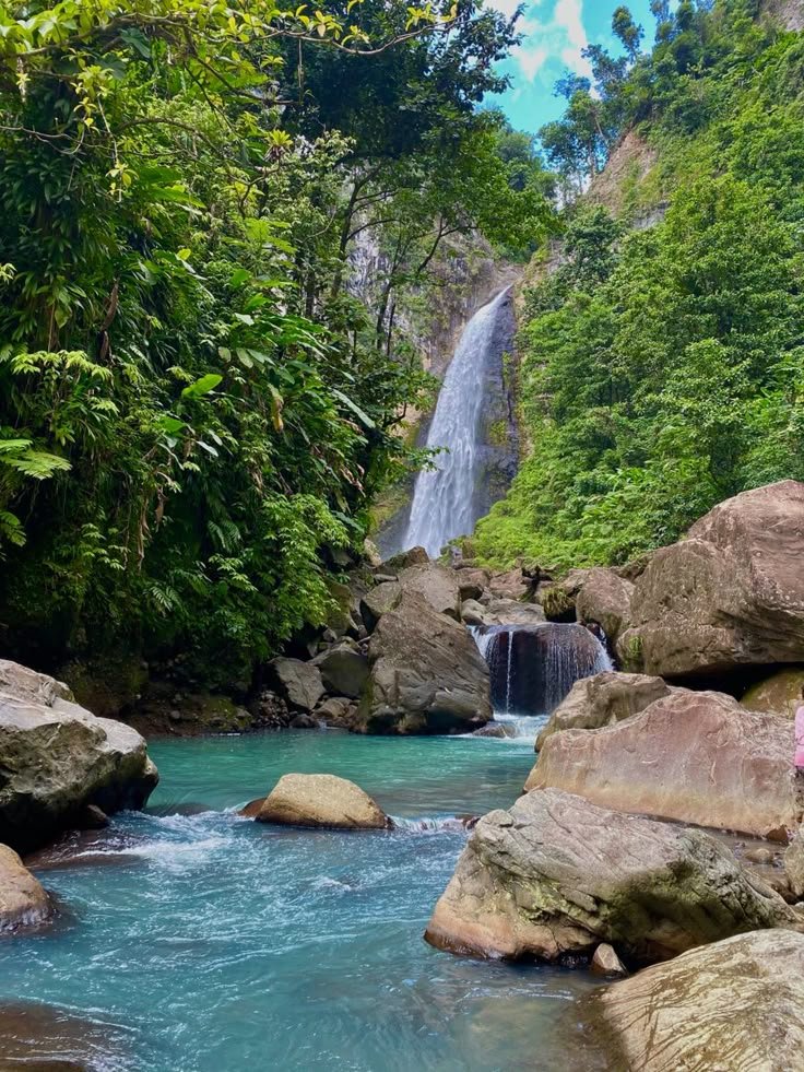 This may contain: a woman standing on rocks in front of a waterfall with blue water and green trees