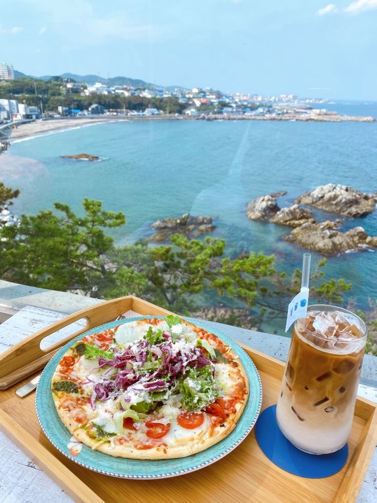 This may contain: a pizza on a tray with a drink in front of the ocean and cityscape