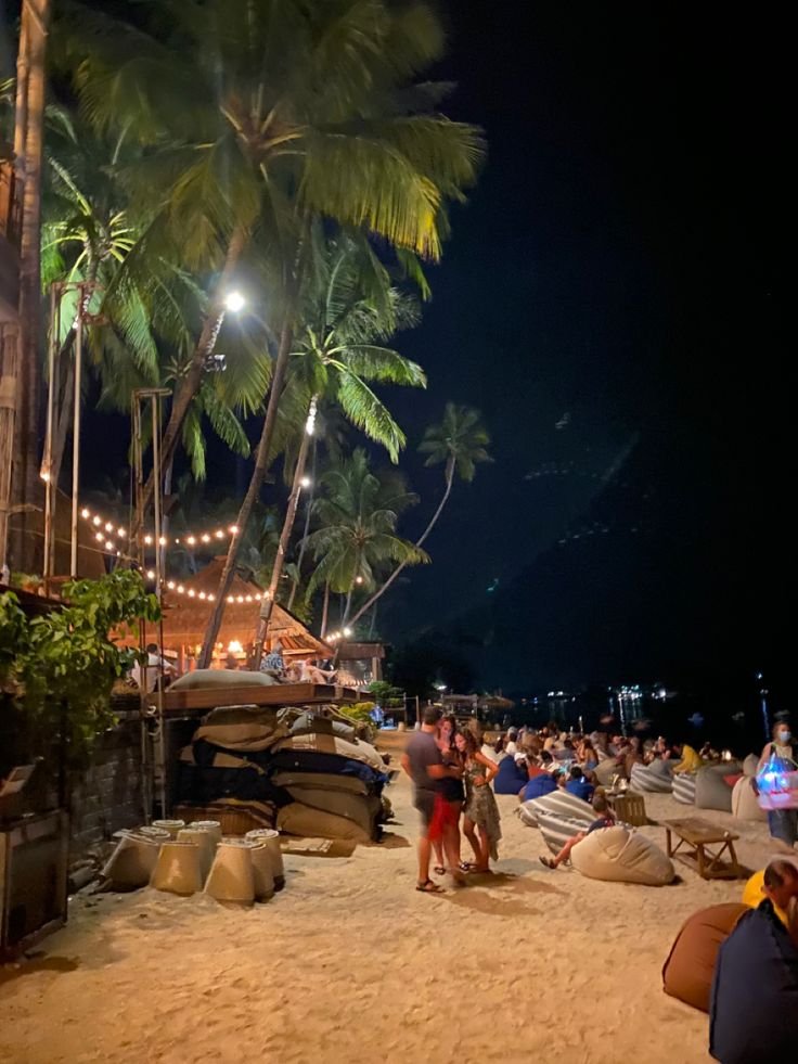 This may contain: people sitting on the beach at night with palm trees and boats in the water behind them