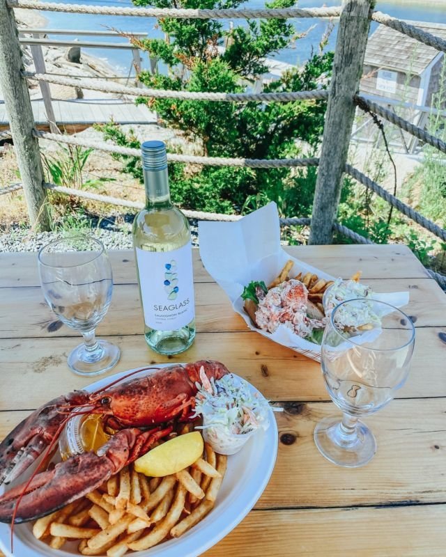 This may contain: lobster, fries and wine on a wooden table with a view of the ocean in the background