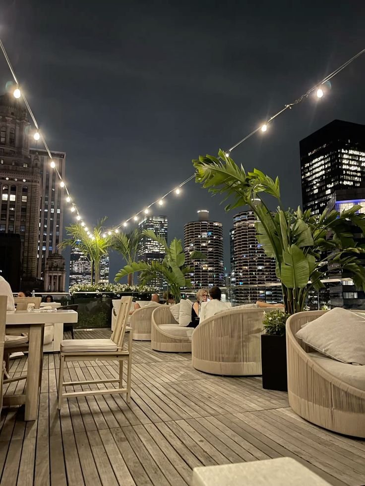 This may contain: an outdoor seating area with lights strung over the city skyline at night, along with chairs and tables