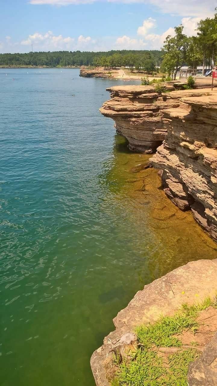 This may contain: a body of water with rocks on the shore and trees in the background, near a cliff