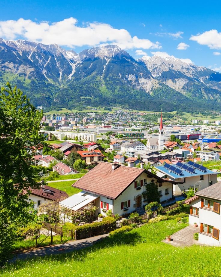 This may contain: a small town with mountains in the background and green grass on the ground, surrounded by trees
