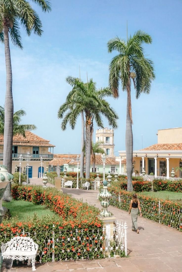 This may contain: a woman walking down a sidewalk next to palm trees and flowers in front of a building