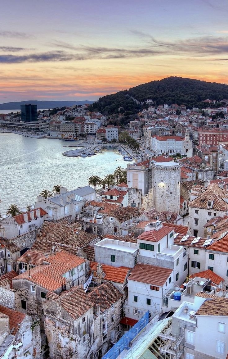 This may contain: an aerial view of a city with red roofs and water in the background at sunset