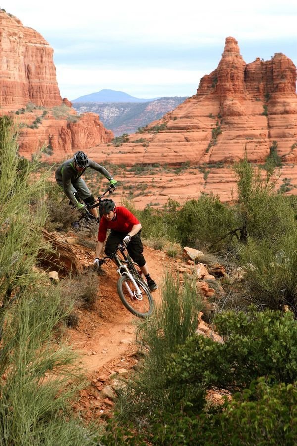 This may contain: two people riding mountain bikes on a trail in the middle of desert area with mountains and trees