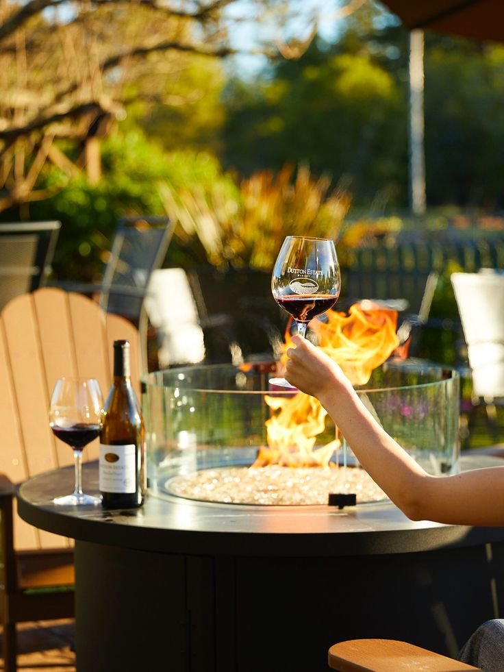 This may contain: two people toasting wine glasses over a fire pit on an outdoor table with chairs