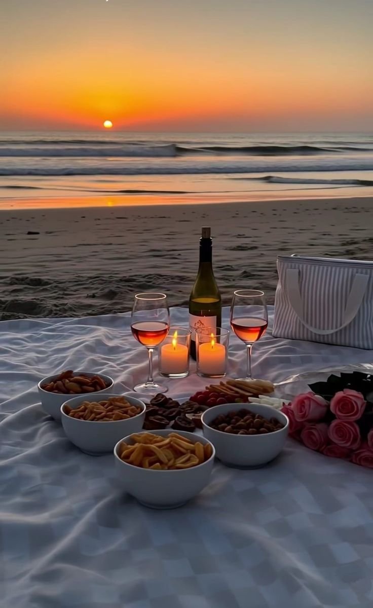 This may contain: two glasses of wine and some food on a table at the beach with sunset in the background