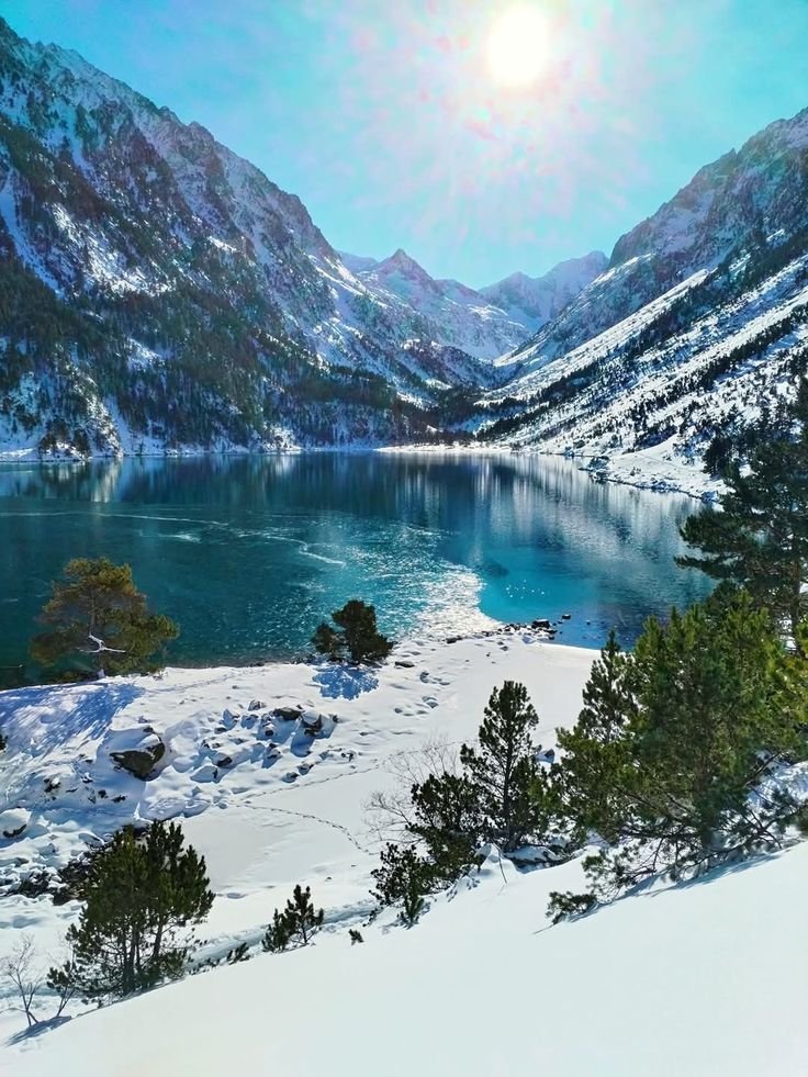 This may contain: the sun shines brightly over a lake surrounded by snow covered mountains and pine trees