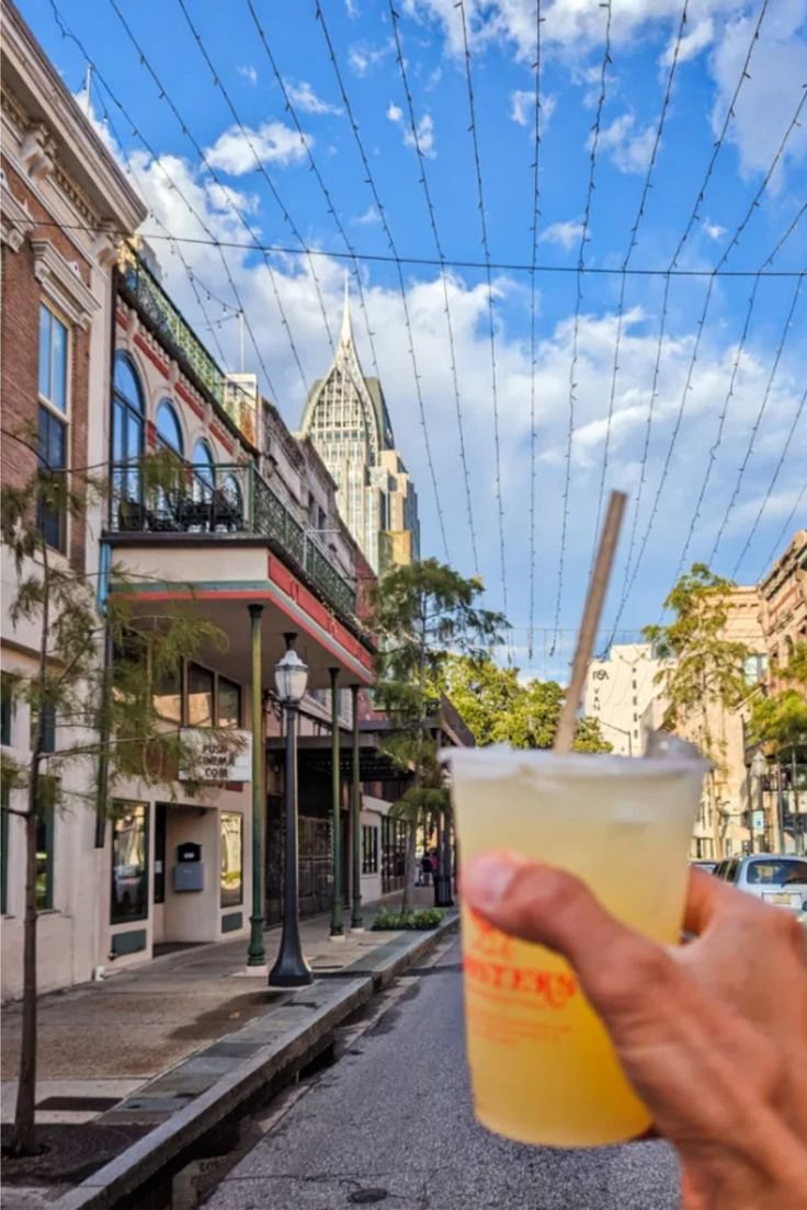 This may contain: a person holding up a drink in front of a building on a street with power lines overhead