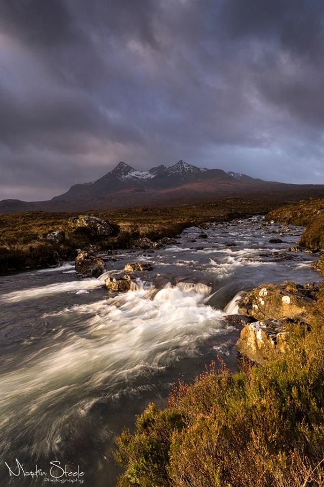 This may contain: a river running through a lush green field under a cloudy sky with mountains in the background