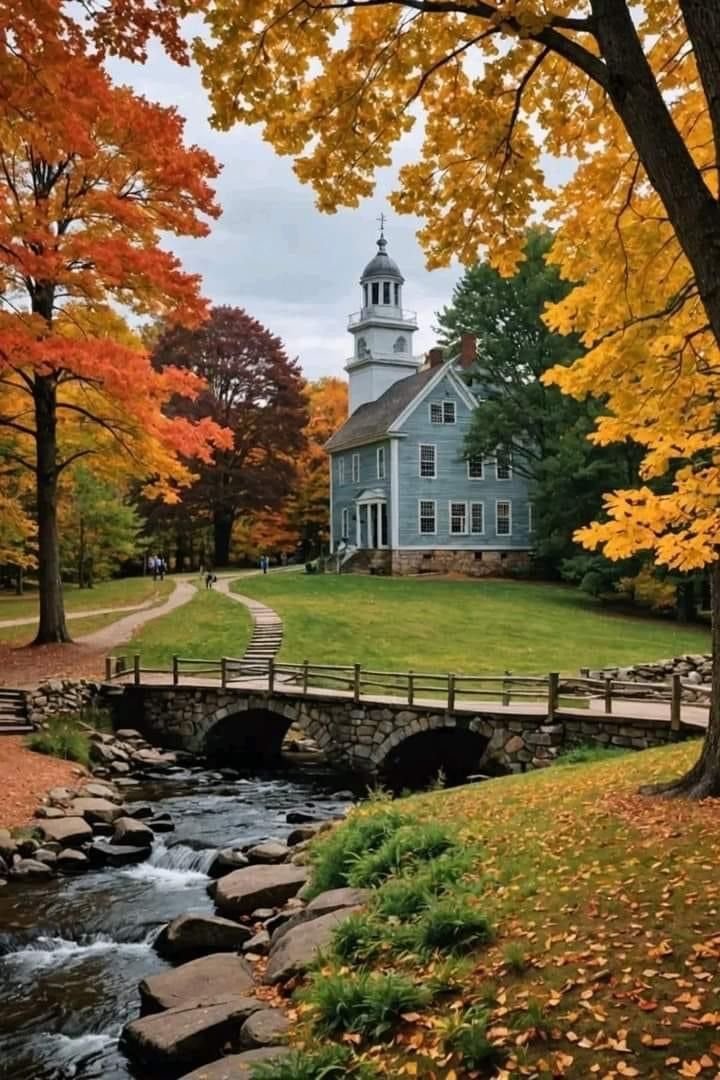 This may contain: a small bridge crosses over a stream in front of a white house with a steeple on top