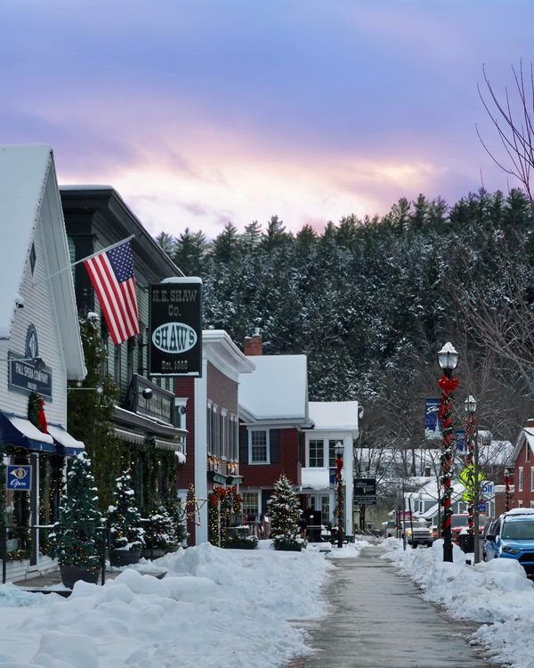 This may contain: a snowy street with cars parked on the side and trees covered in snow at dusk