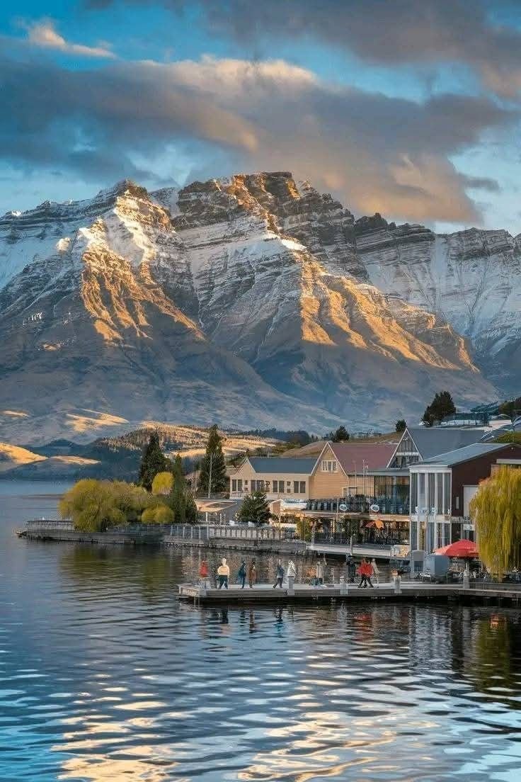 This may contain: the mountains are covered in snow as people sit on a dock near some buildings and trees
