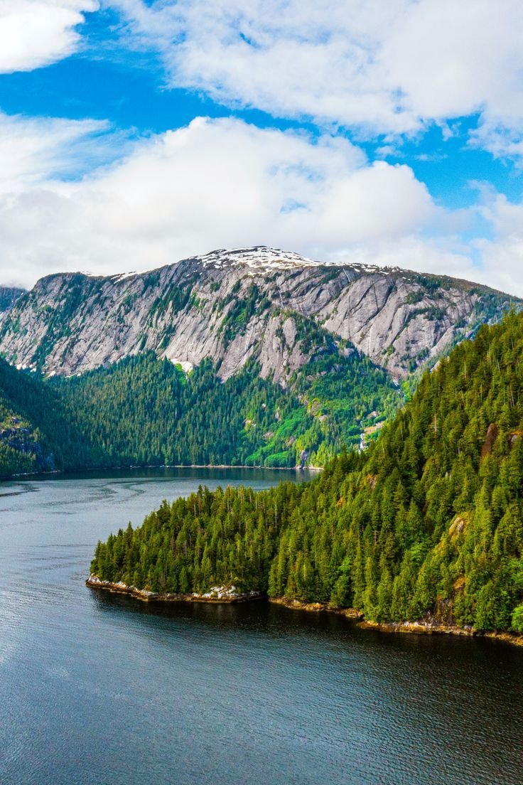 This may contain: an island in the middle of a body of water surrounded by trees and mountains under a blue sky with white clouds